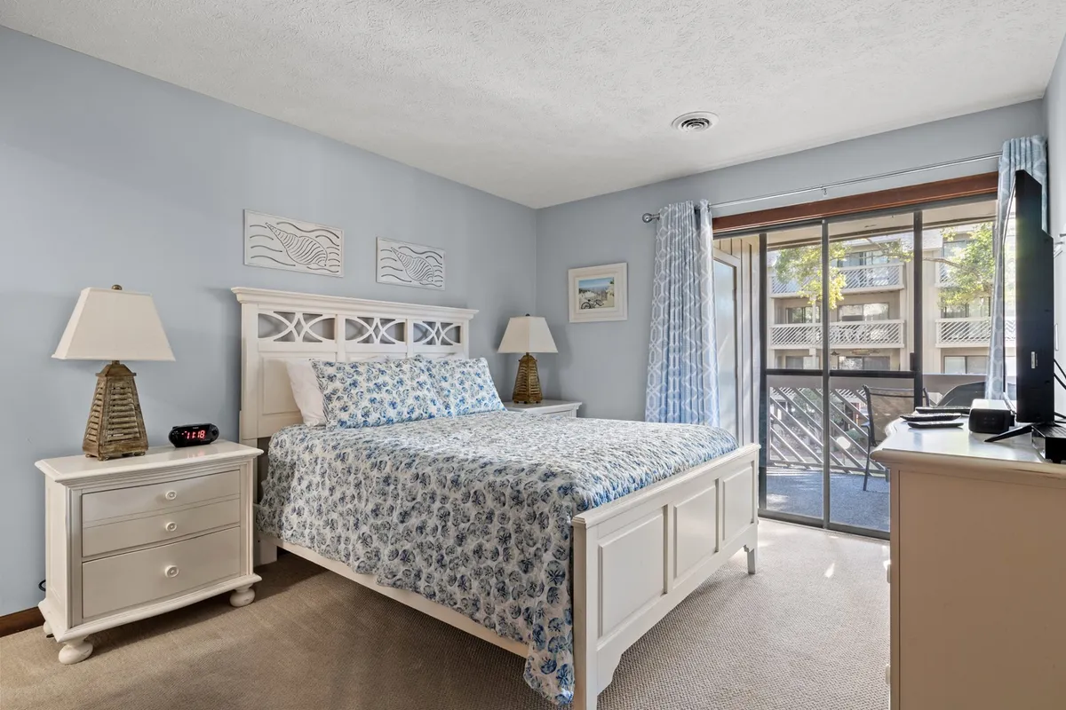 This image features a bedroom with a bed dressed in floral bedding, a large window with sheer curtains allowing soft, natural light to fill the room, and a nightstand with a lamp. The room exudes a peaceful and serene atmosphere, ideal for restful retreats.