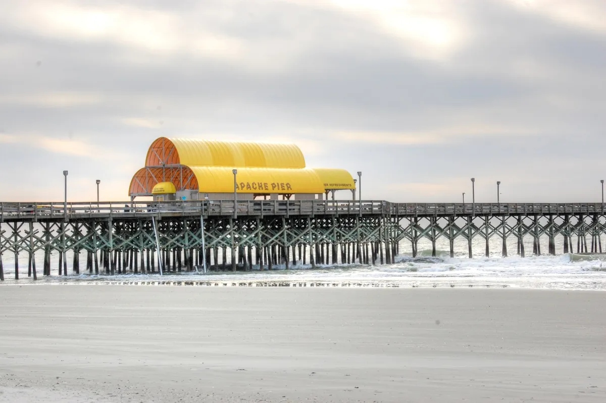 This image showcases the iconic Apache Pier extending over the ocean, with gentle waves rolling onto the sandy beach. The scene features a tranquil, early-morning or sunset ambiance, inviting visitors to take a leisurely stroll or enjoy the soothing sights and sounds of the water.