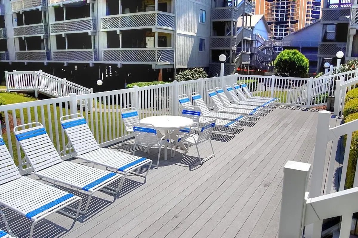 A sun-drenched exterior pool area with inviting loungers, a circular table, and white railings, overlooking a serene setting. This space promises relaxation, whether sunbathing, reading, or unwinding after a day at the beach.