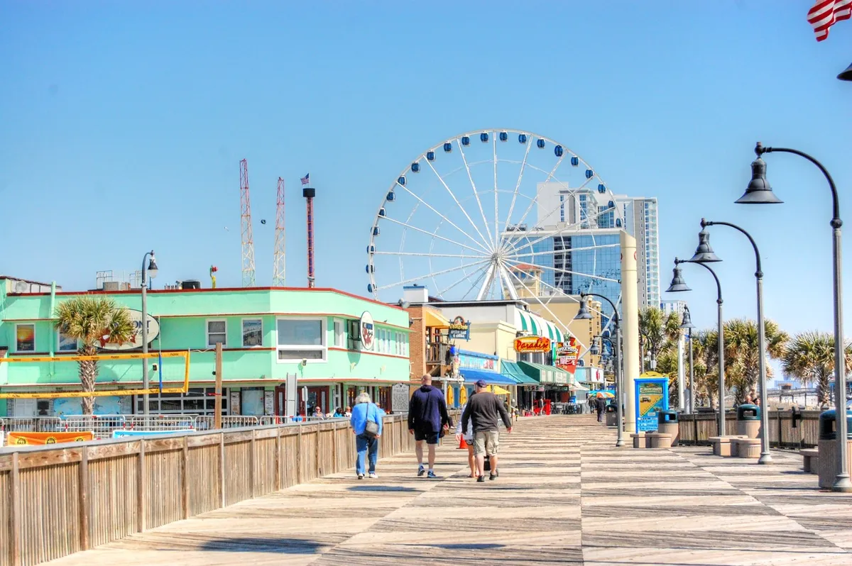 The iconic Myrtle Beach SkyWheel rises above colorful boardwalk buildings, with people strolling below. The lively scene captures the excitement and variety of local attractions available to guests.