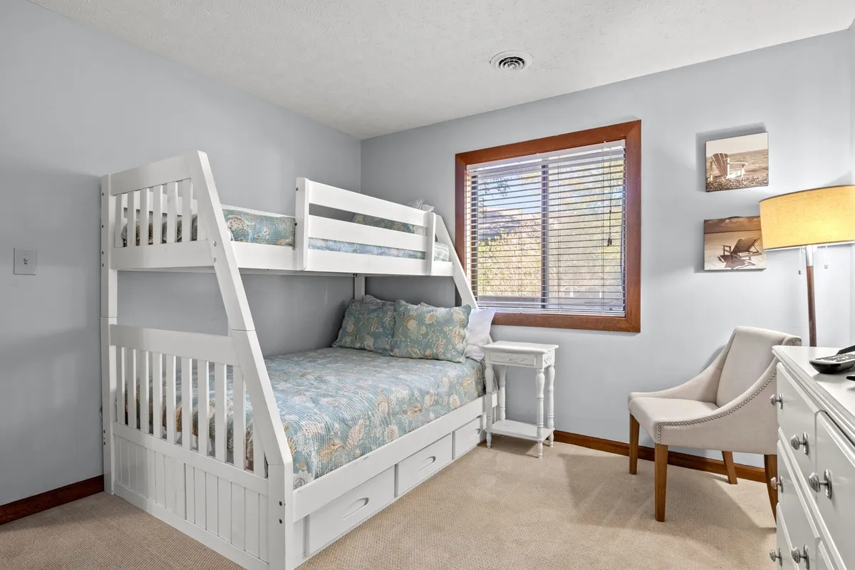 This image showcases a second bedroom with a bunk bed (pyramid style), a window with blinds streaming in morning light, a nightstand, an armchair, and a lamp. The setup feels both playful and practical, catering to families or groups.