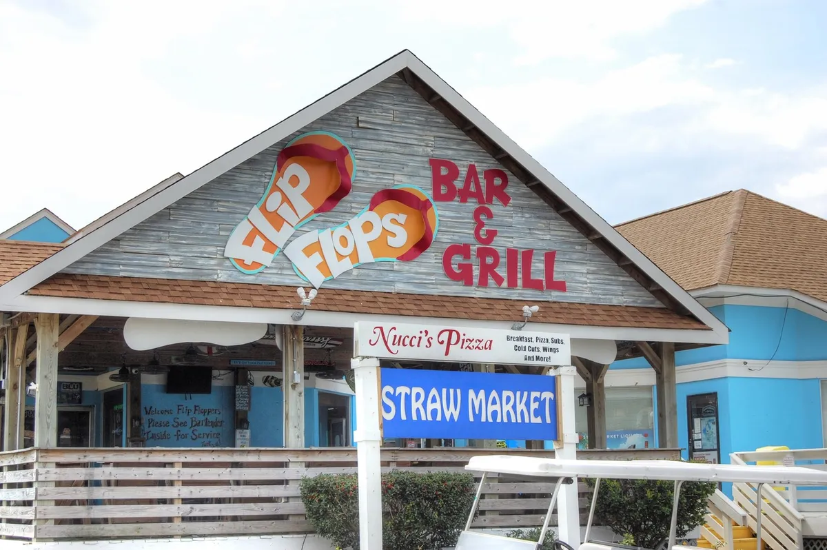 A colorful exterior shot featuring the welcoming signs for Flip Flops Bar & Grill, Nucci's Pizza, and the Straw Market, hinting at a lively local dining and entertainment scene. The area gives off a laid-back beach-town vibe, perfect for spontaneous outings and casual evenings.