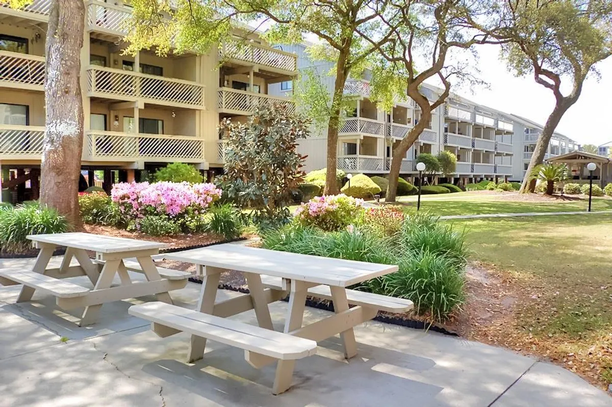 An exterior view featuring a cheerful picnic area set among lush flower beds and mature trees, with building facades in the background. A picnic table provides a perfect spot for outdoor meals, while the vibrant landscaping invites groups to gather, relax, and create memories together.