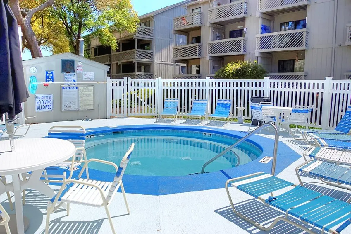 An exterior view of a hot tub area with lounge chairs, a fenced space, and a poolside table. The ambiance is relaxed and ideal for sunbathing, poolside socializing, or enjoying a dip in the hot tub.