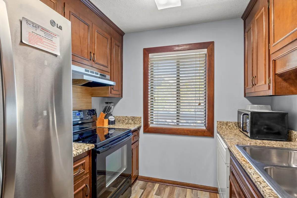 A welcoming kitchen scene featuring a stainless steel refrigerator, granite countertops, a window with blinds allowing in natural light, and a full suite of modern kitchen appliances. The countertops are clear and inviting, suggesting readiness for meal prep or casual conversation.