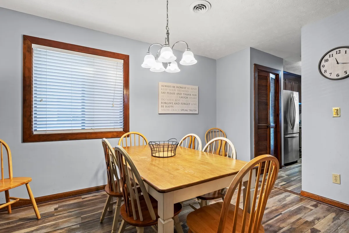 A bright dining area with a stylish table set for meals, a modern light fixture above, wall art, a clock, and a window with blinds allowing natural light to filter in. The space feels warm and ready for family meals or friendly gatherings.