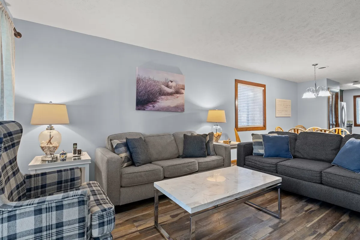 An open living space with a plaid armchair, grey sofas, a marble coffee table, and wall art, set against a backdrop of natural light streaming in. The layout is conducive to group activities like board games, conversations, and relaxed reading, creating an inviting and communal vibe.