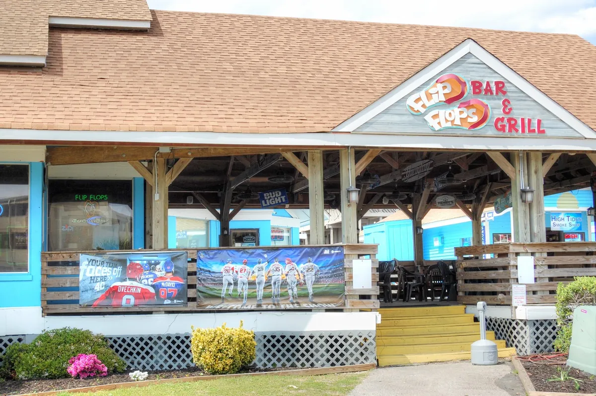 A lively exterior scene showing the colorful signage of 'Flip Flops Bar & Grill,' highlighted by festive banners and an inviting outdoor seating area. The space is clearly designed for casual dining, socializing, and enjoying themed events or live entertainment—perfect for groups seeking local fun.