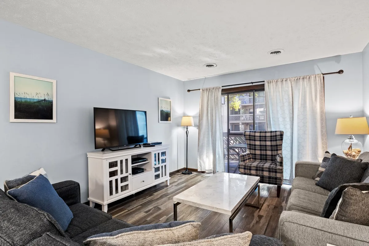 A comfortable living area featuring a plush sofa, coffee table, flat-screen television, stylish lamp, and curtains framing a sunlit window. The arrangement invites relaxation and social gatherings with ample seating and a soft, welcoming mood.