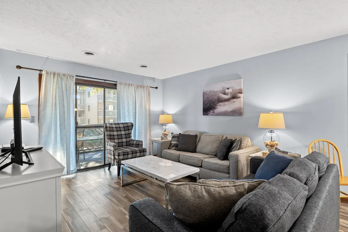 The image showcases a cozy living room featuring a plush sofa, stylish armchair, and a central coffee table, all bathed in natural light streaming through large curtains. Subtle coastal artwork and comfortable furnishings invite relaxation, with the balcony just beyond—perfect for sunset views or unwinding after a day at the beach.