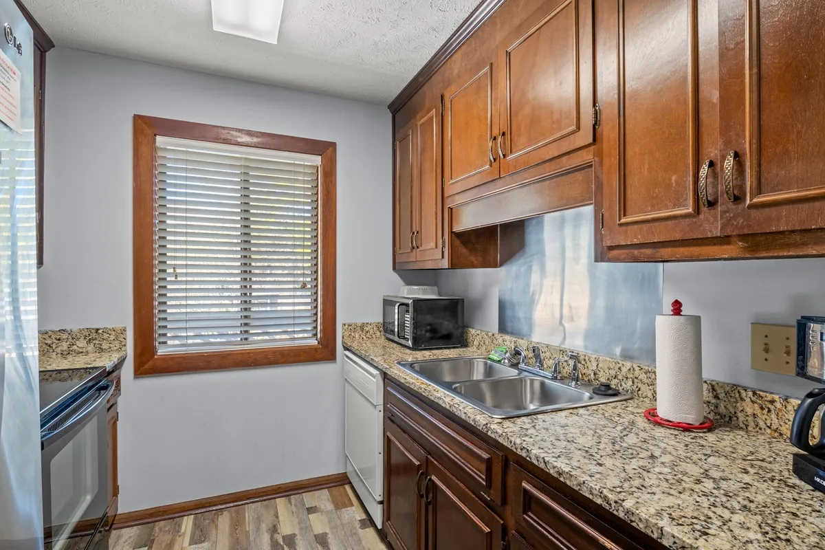 A cozy kitchen corner with granite countertops, warm wooden cabinets, a sunlit window with blinds, and visible kitchen appliances. The setting feels homey and intimate, perfect for family meal prep or holiday baking.