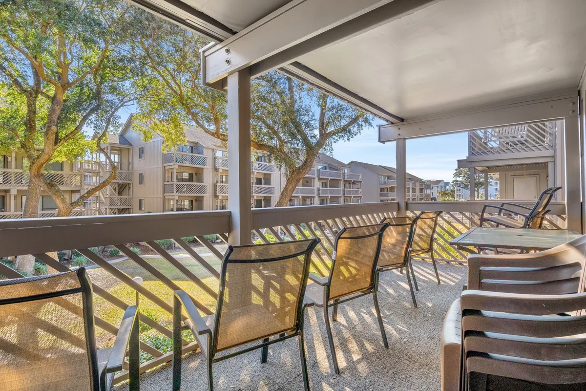 This exterior image captures a private balcony with a comfortable sitting area, surrounded by trees and overlooking adjacent buildings. The scene is inviting, showing a perfect spot for morning coffee, reading, or enjoying sunset cocktails while being just steps from the ocean.